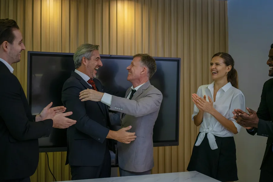 Zwei Unternehmer gratulieren sich nach der gelungenen Übergabe, das Team applaudiert